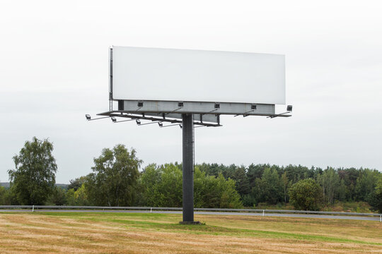 Large commercial megaboard, billboard standing in a field next to road and trees on a skyline with empty blank space gray sky background.