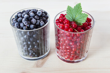 Two glasses full of summer berries - redcurrants and blueberries with decorative green leaf on wooden table surface.