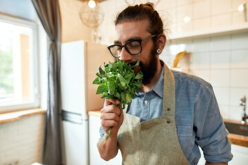 Close up of young man, Italian cook in apron smelling basil leaves while getting ready to prepare a meal, standing in the kitchen. Cooking at home concept