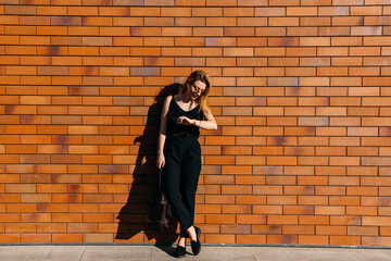 Fototapeta na wymiar Young businesswoman, standing on a brick wall background outdoors, looking to her watch. Business concept.