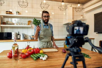 Young man, Italian cook in apron looking at camera, holding basil, filming himself for culinary...