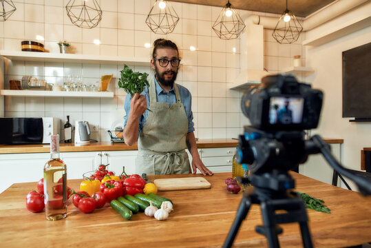 Young man, Italian cook in apron looking at camera, holding basil, filming himself for culinary blog while preparing healthy meal with vegetables in the kitchen