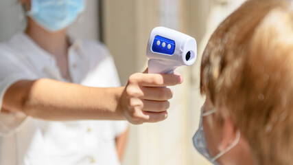 Teacher with facemask scanning a kid with digital thermometer, measuring body temperature before lessons. New normal, back to school during coronavirus pandemic.
