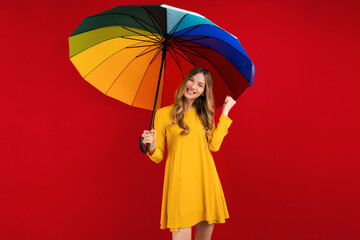 Happy young woman with a rainbow umbrella, shows a winning gesture on a red background