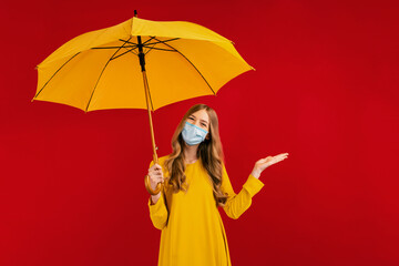 Happy young woman in a yellow dress and a medical protective mask on her face, with an umbrella in her hands, points to the empty space, on a red background