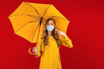 young woman in a medical protective mask on her face, shows a finger on the mask, with an umbrella in her hands, on a red background