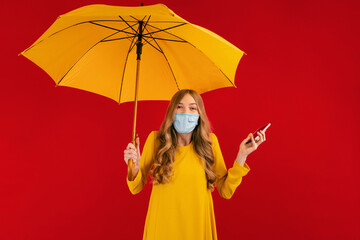 Shocked young woman in a protective mask on her face, with an umbrella in her hands and a mobile phone, on a red background