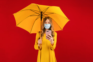 Happy young woman in a protective mask on her face, in a yellow dress, with an umbrella in her hands, with a mobile phone, on a red background