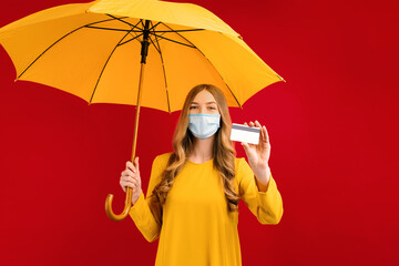 Happy young woman in a medical mask on her face, with an umbrella and a credit card in her hands, on a red background