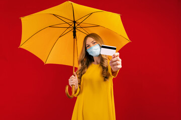 Happy young woman in a medical mask on her face, with an umbrella and a credit card in her hands, on a red background