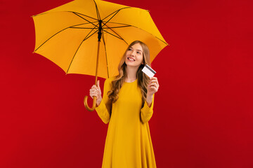 Pensive beautiful young woman with umbrella and credit card in hand, on a red background