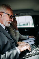 Good looking senior business man and his young woman colleague or coworker sitting on backseat in luxury car. They talking, smiling and using laptop and smart phones. 