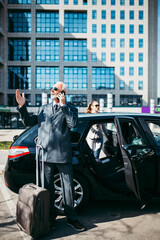 Good looking senior business man and his young woman colleague or coworker standing by the car. He talking on smartphone, holds a suitcase and prepares to go on a business trip. 
