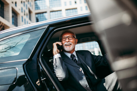 Good Looking Senior Business Man Sitting On Backseat In Luxury Car. He Opens Car Doors And Going Or Stepping Out. Big Business Building In Background. Transportation In Corporate Business Concept.