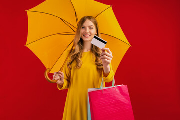 Happy beautiful young woman with umbrella holding shopping bags and credit card on red background, shopping concept, autumn