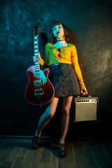 Grunge style young hipster woman with curly hair holding red guitar and amp in neon lights. 90s...