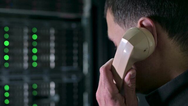 A man calls on an old phone against the backdrop of a modern server cabinet. A comic image of the mixing of eras of technology. Obsolescence of technology