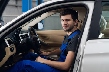 Car service worker in overalls sitting in a car in the front seat and looking at the camera