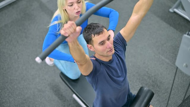 Slim guy performs an exercise on an exercise machine under the supervision of a fitness club trainer, top view