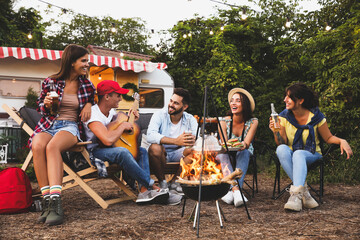 Happy friends sitting near bonfire. Camping season - obrazy, fototapety, plakaty