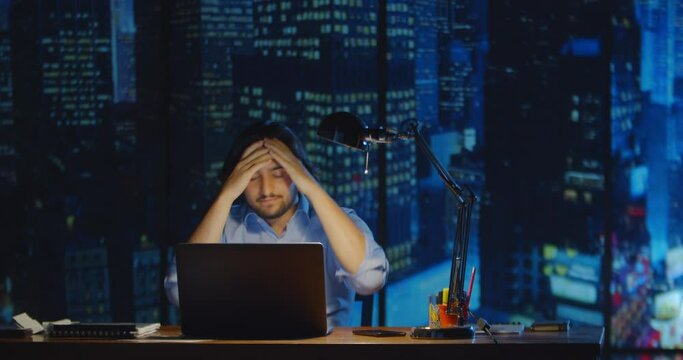  Frustrated businessman at night office after a difficult day at work. The trader hits the table because of the lost money on the exchange. Business and finance in a big city