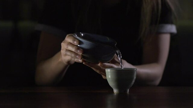 Young woman bartender pours sake from blue ceramic Katakuchi into green Guinomi (sake cup) - Slow motion