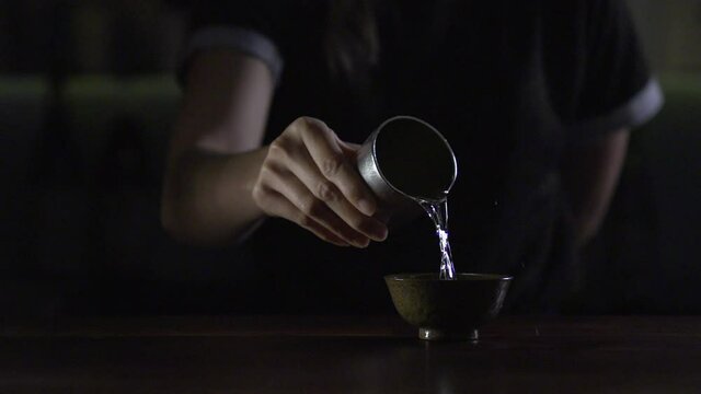 Young woman bartender pours sake from silver sake pitcher into dark green Guinomi (sake cup) - Slow motion