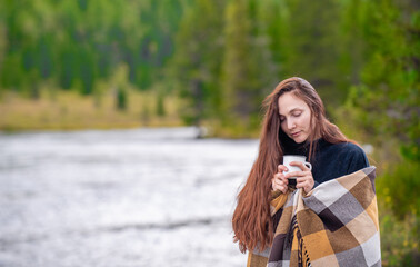 Young woman wrapped in a plaid drinks tea from a mug outdoors