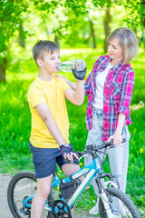 Obraz premium Young boy drinks water from a bottle while riding a bicycle. Mom cares about her son's health on a hot summer day