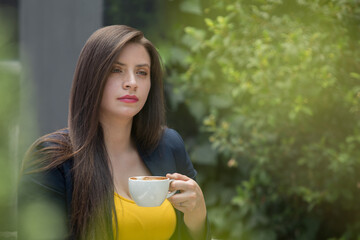 young white-skinned woman with long straight hair, holding in her hand a white cup, wears a yellow blouse and a blue jacket, in the background is a bush