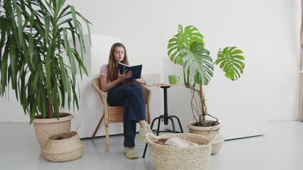 Beautiful woman reading a book in her living room at home. Relaxed young woman sitting near house plants