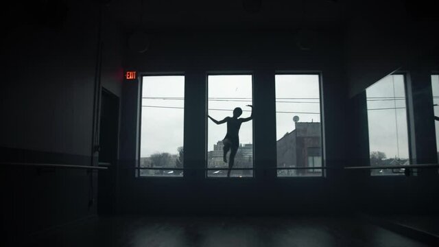 Dancer on window sill dancing in large dark moody dance studio with windows in the background in silhouette