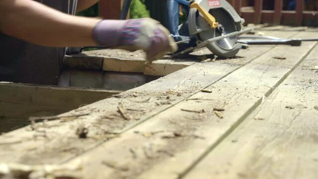 Gloved man uses crow bar to lift deck board and then uses hammer to remove nail