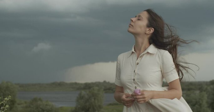 Headshot portrait of girl breathing fresh windy air on rainy stormy sky background. Serious woman standing with blowing fluttering hair outdoor slow motion copy text space. Environment protection
