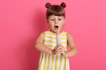 Surprised female child wearing white and yellow summer dress holding water ice cream, keeps mouth widely opened, being astonished, standing isolated over rosy wall.