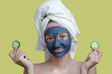 A young girl with a white towel on her head, a cosmetic clay mask on her face, holding cucumber slices on a yellow isolated background.Concept of Spa treatments, facial care