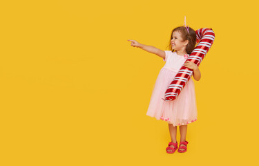 A cute little girl child in a fancy dress and a Santa hat holds an inflatable shape of a candy cane on a yellow background. 2021 new year holidays concept. points to the left