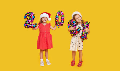 Cheerful children in Santa hats holding colorfull numbers 2021  on yellow background. Happy New Year party with balloons.