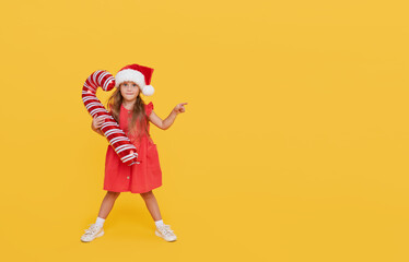 A cute little girl child in a fancy dress and a Santa hat holds an inflatable shape of a candy cane on a yellow background. 2021 new year holidays concept. points to the right