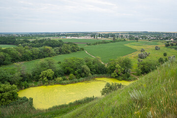 Fototapeta premium Wonderful rural landscape. Valley view with green lake, fields and village on blue sky background