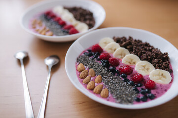 Smoothie bowl with fresh raspberry, blueberry, chia seeds, almonds, banana, and chocolate flakes. Morning breakfast on wooden table. Two ptales fith smoothie bows