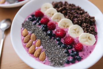 Smoothie bowl with fresh raspberry, blueberry, chia seeds, almonds, banana, and chocolate flakes. Morning breakfast on wooden table. Close up