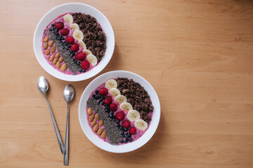 Smoothie bowl with fresh raspberry, blueberry, chia seeds, almonds, banana, and chocolate flakes. Morning breakfast on wooden table. Top view of two plates. vegan food