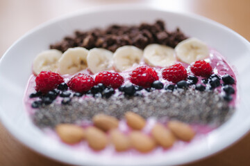 Smoothie bowl with fresh raspberry, blueberry, chia seeds, almonds, banana, and chocolate flakes. Morning breakfast on wooden table. Close up