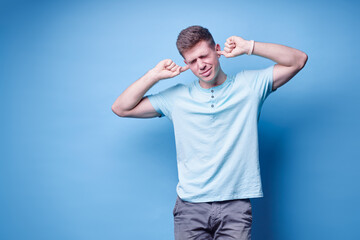 To loud! Young man closes his ears with fingers refusing to listen to something. Blue background.