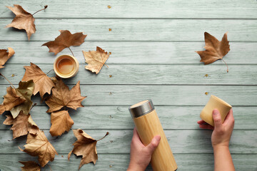 Zero waste eco friendly insulated bamboo flask with bamboo cups. Trendy flat lay with hands holding the flask and natural bamboo cup on aged blue mint wooden table with Autumn sycamore leaves.