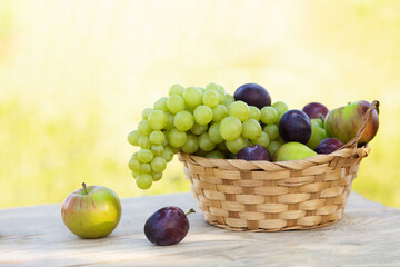 a basket with apples, plums and grapes on a light wooden table on a yellow nature background