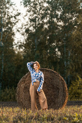 Beautiful modern  young woman wearing in beige trousers and a plaid shirt posing against   a haystack in field. Trendy teenage girl in autumn outdoors.