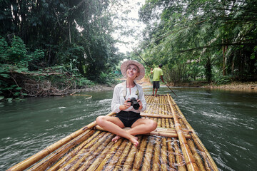 Fototapeta na wymiar Traveling by Thailand. Pretty young woman enjoying view sailing jungle river on traditional bamboo raft.