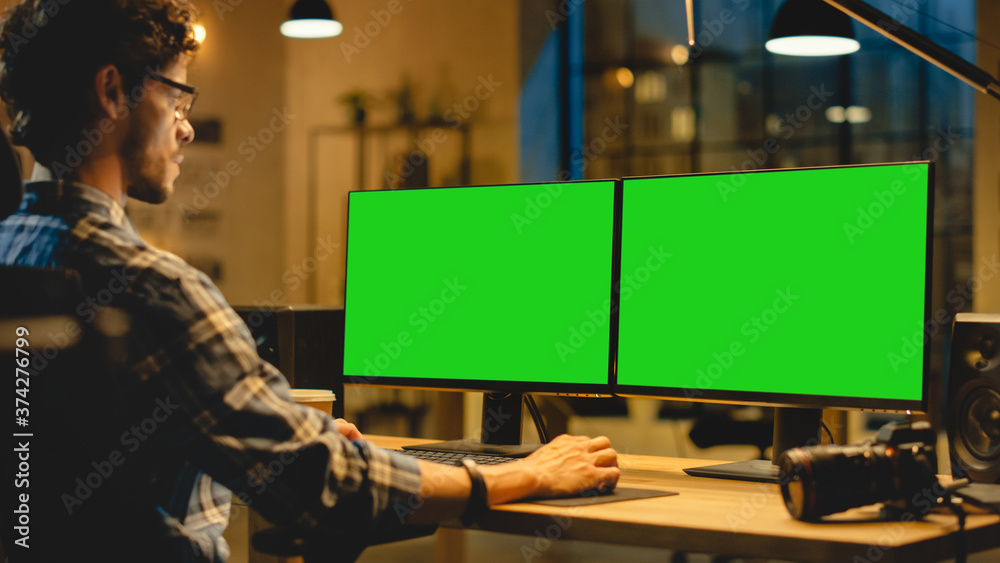 Poster over the shoulder: creative designer sitting at his desk uses desktop computer with two green mock-u